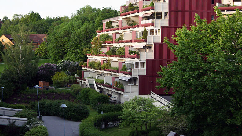 Landskapsbilde av rødmalt terraseblokk med hvite balkonger og med trær og busker rundt