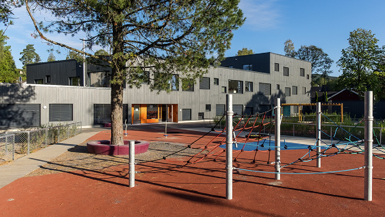 Moderne, grått barnehagebygg over flere etasjer med klatrestativ i forgrunnen