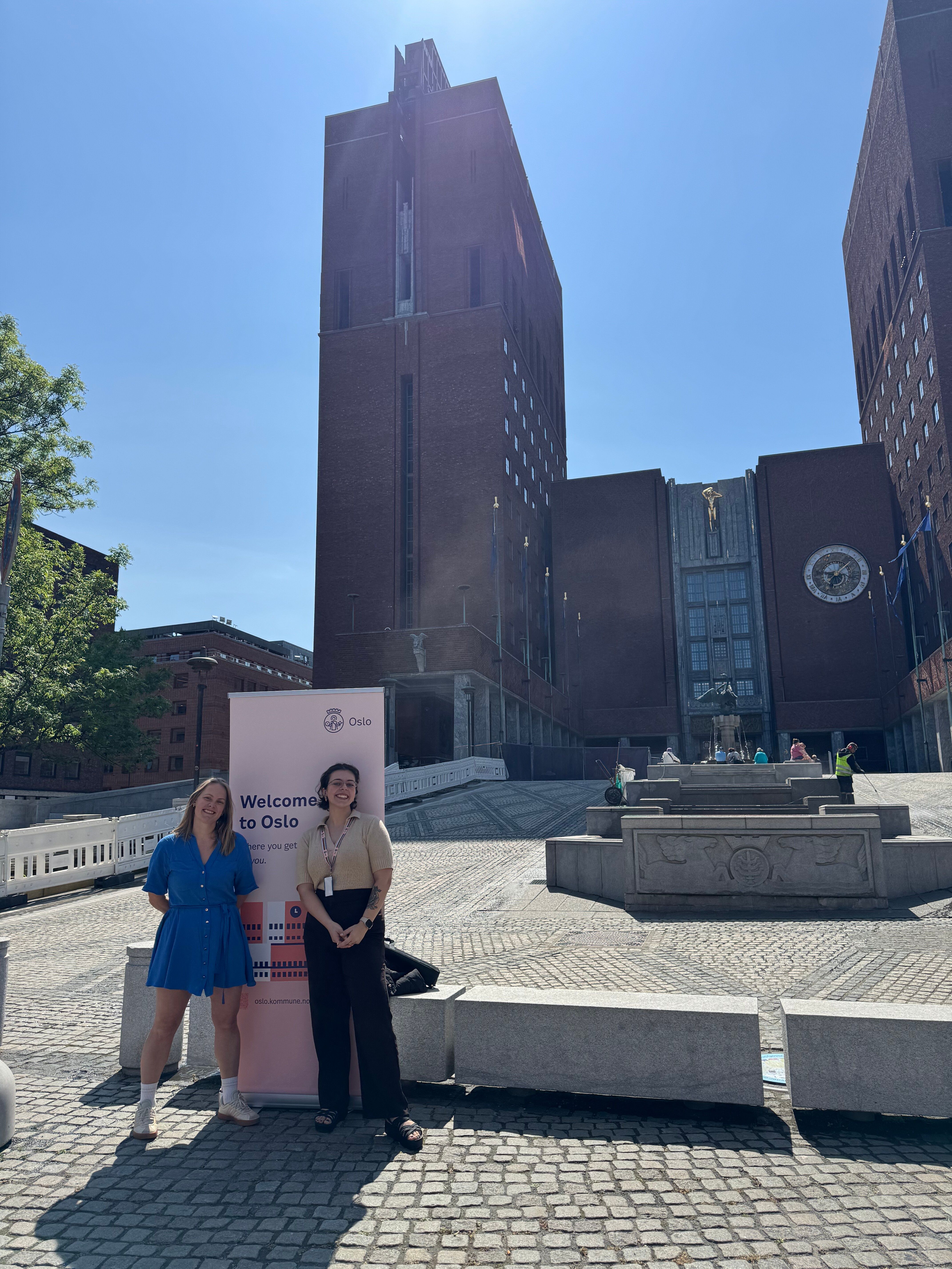 Two women standing in front of the Oslo radhuset with a roll up sign that says Welcome to Oslo