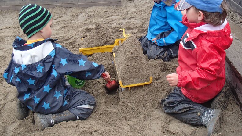 tre barn med regntøy som leker i sandkassen