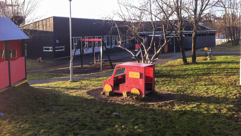 Uteområde med rødmalt fastmontert stor lekebil på gress, husker, trær og i bakgrunnen en svartmalt barnehage