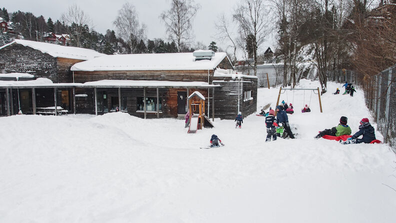 Uteområde om vinteren, med sklie, huske og med barn som leker i snøen, mørk gråmalt barnehage i bagrunnen