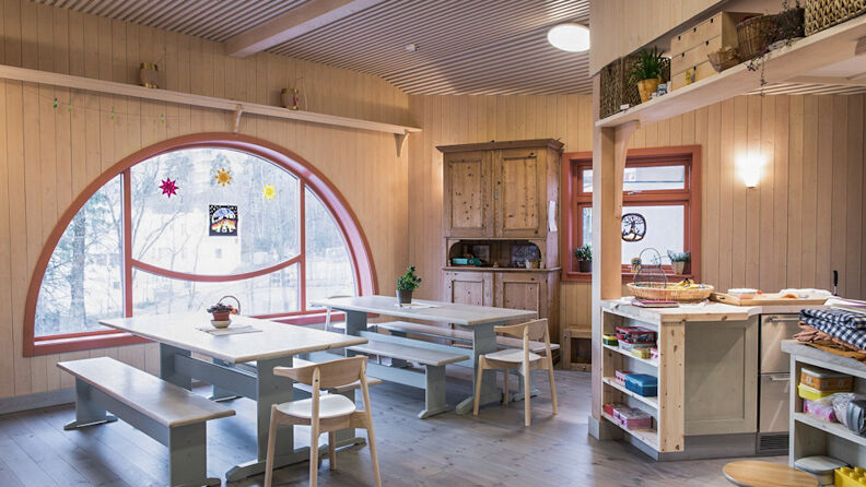 Hvitmalt rom med rødmalte lister rundt vinduene, bord, stoler, hyller og barnetegninger i vinduet